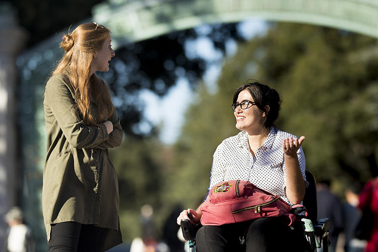 Two students talking in front of Sather Gate. One of the students is a wheelchair user.