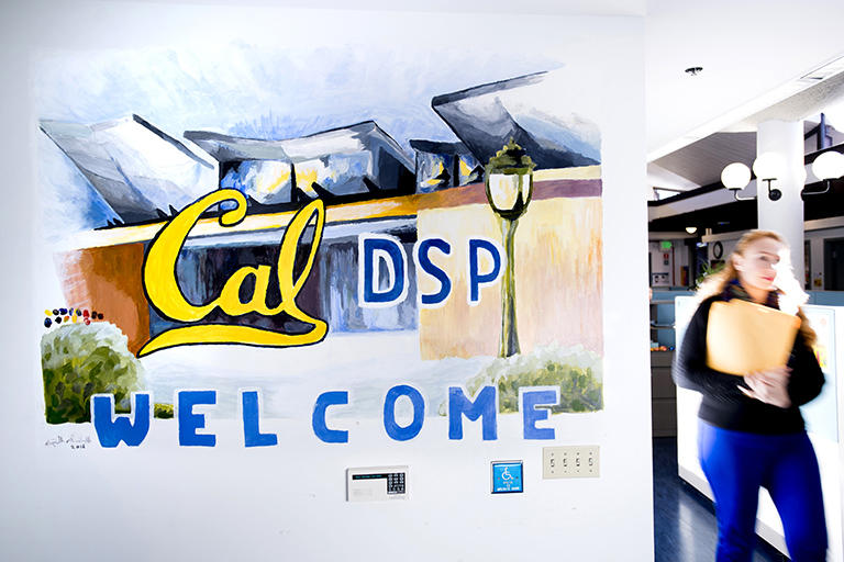 A staff member walking to the right of the mural painted at DSP. The mural reads "Cal DSP, Welcome" and has a painting of the Cesar Chavez Building in its background. 
