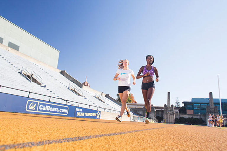 Two students running side by side on a track, with a set of bleachers in the background. 