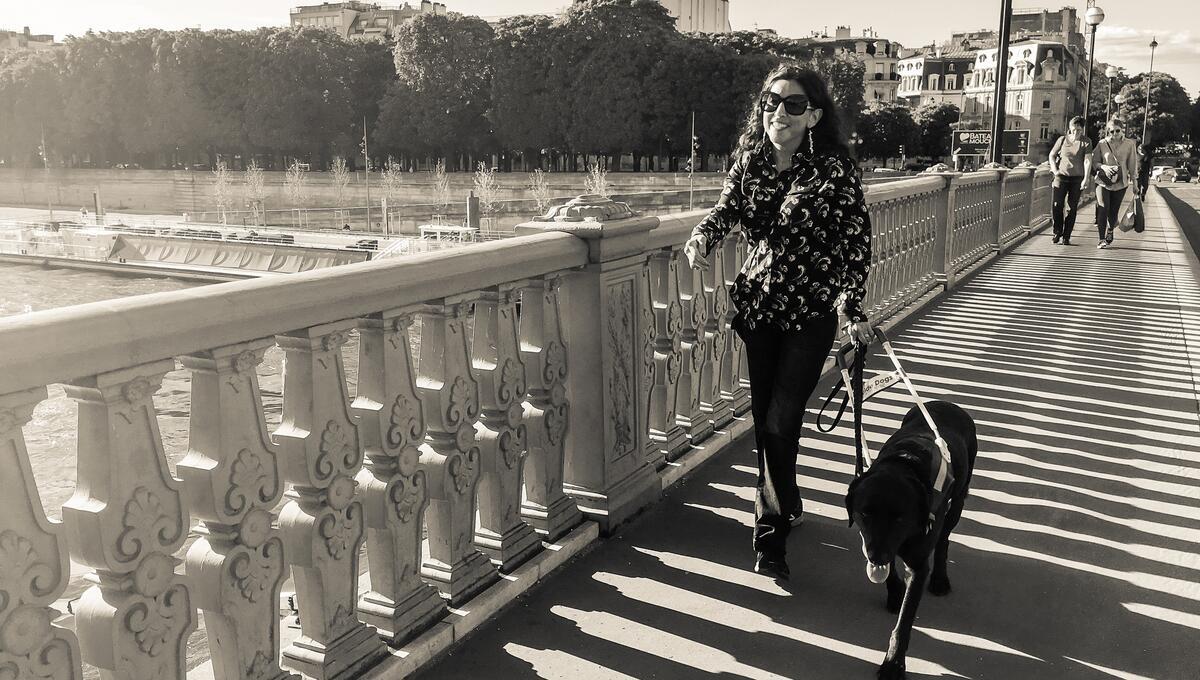 Photo is black and white and shows the photogarpher's wife, Melissa, smiling, wearing sunglasses, walking across a bridge with her guide dog, Camry, with water and buildings in the background.